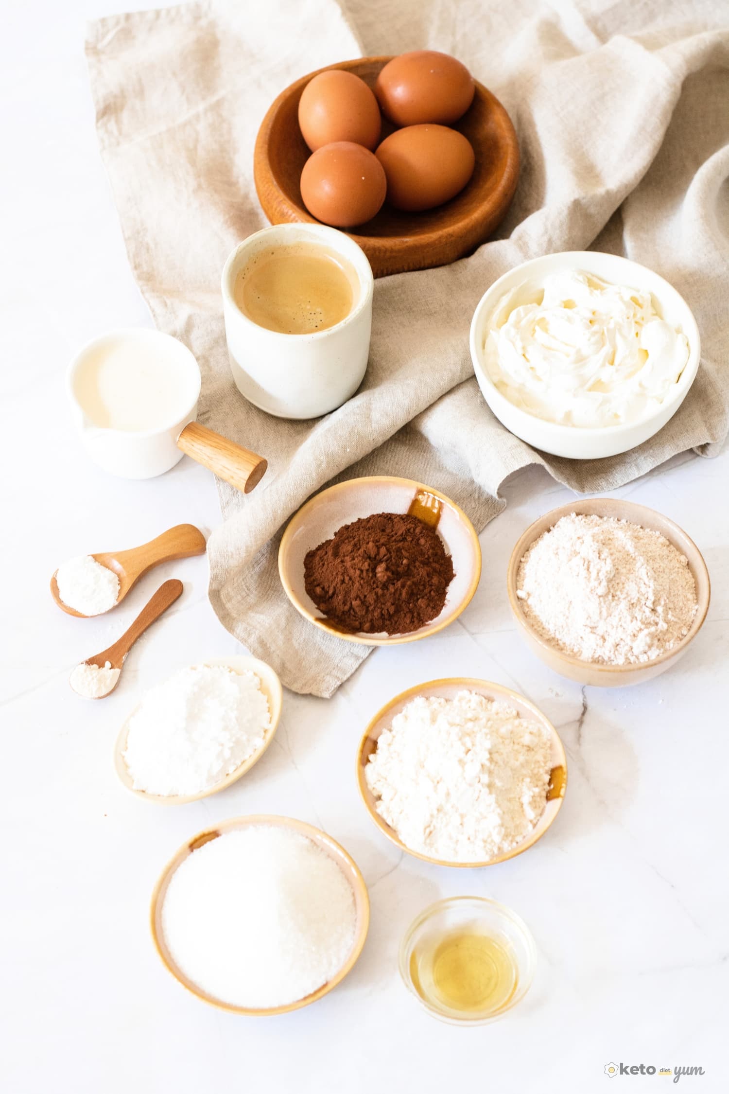 Ingredients for a keto tiramisu recipe laid out on a white surface, including eggs, almond flour, cocoa powder, and cream.